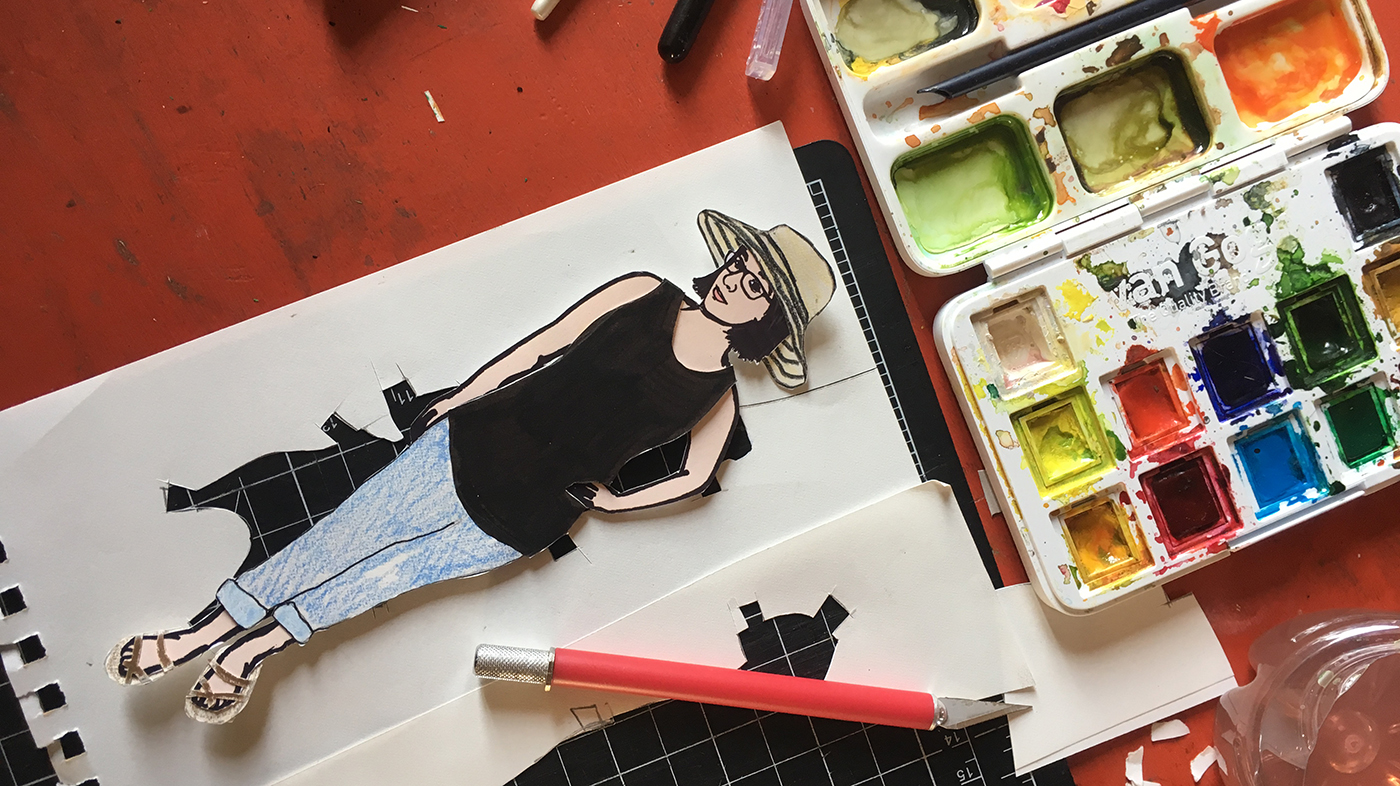 paper doll of the artist on worktable with paint set, exacto knife, and cutting mat.
