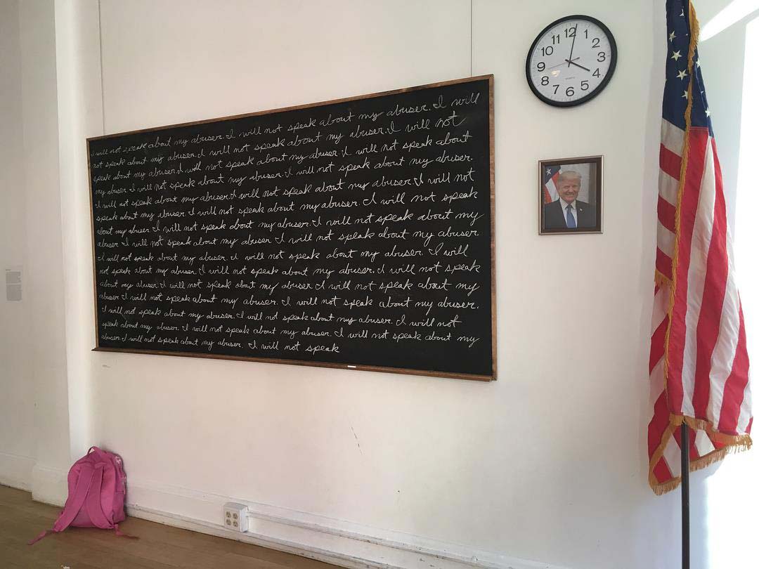 photo of installation which includes from right to left and small pink backpack on the floor, large chalkboard with "I will not speak about my abuser" written over and over agian, school clock and photo of President Trump on wall, and large standing American flag.