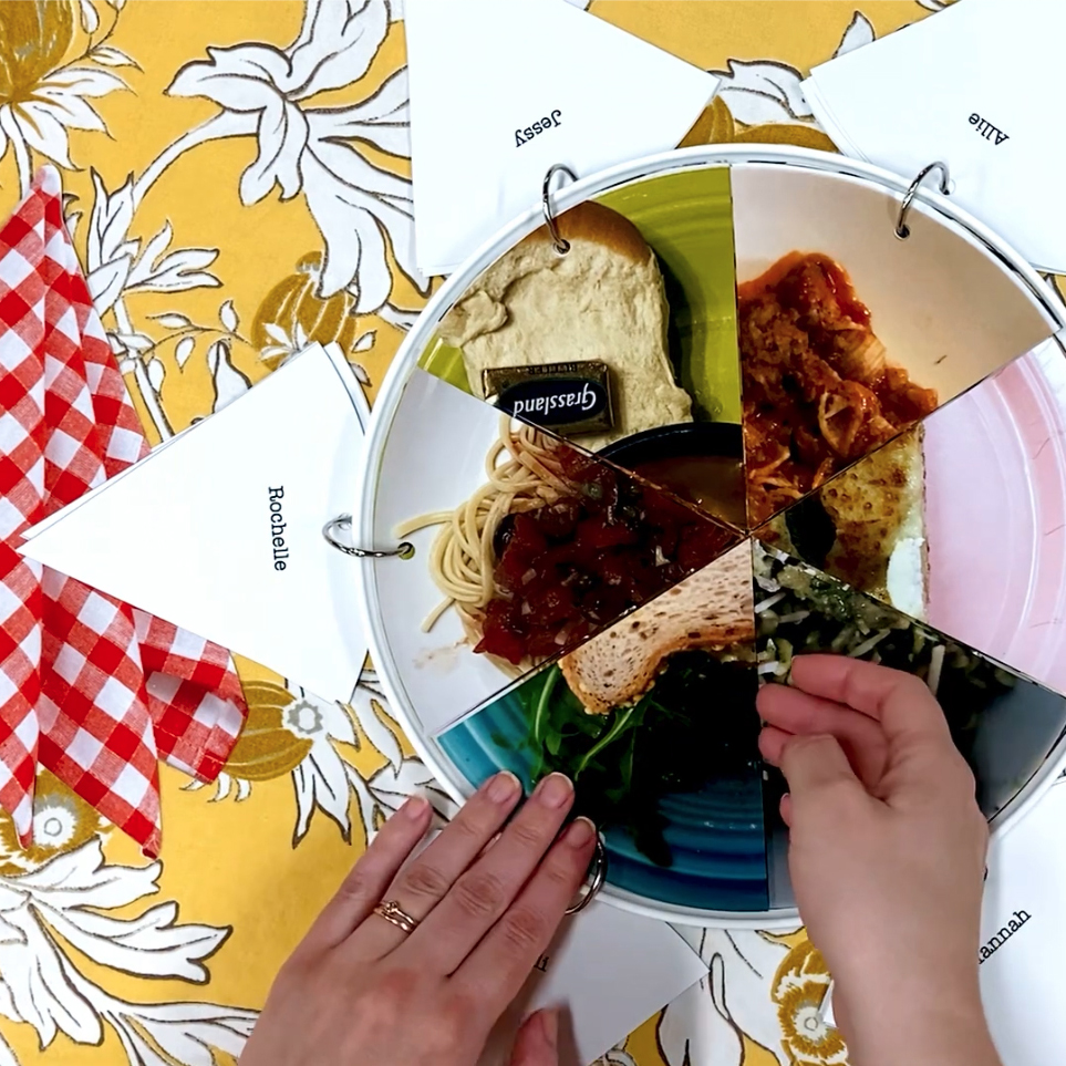 Top down view of hands opening wedge-shaped pages in a circular book depicting food on dinner plates. Link to Dinner Unparty: a potluck