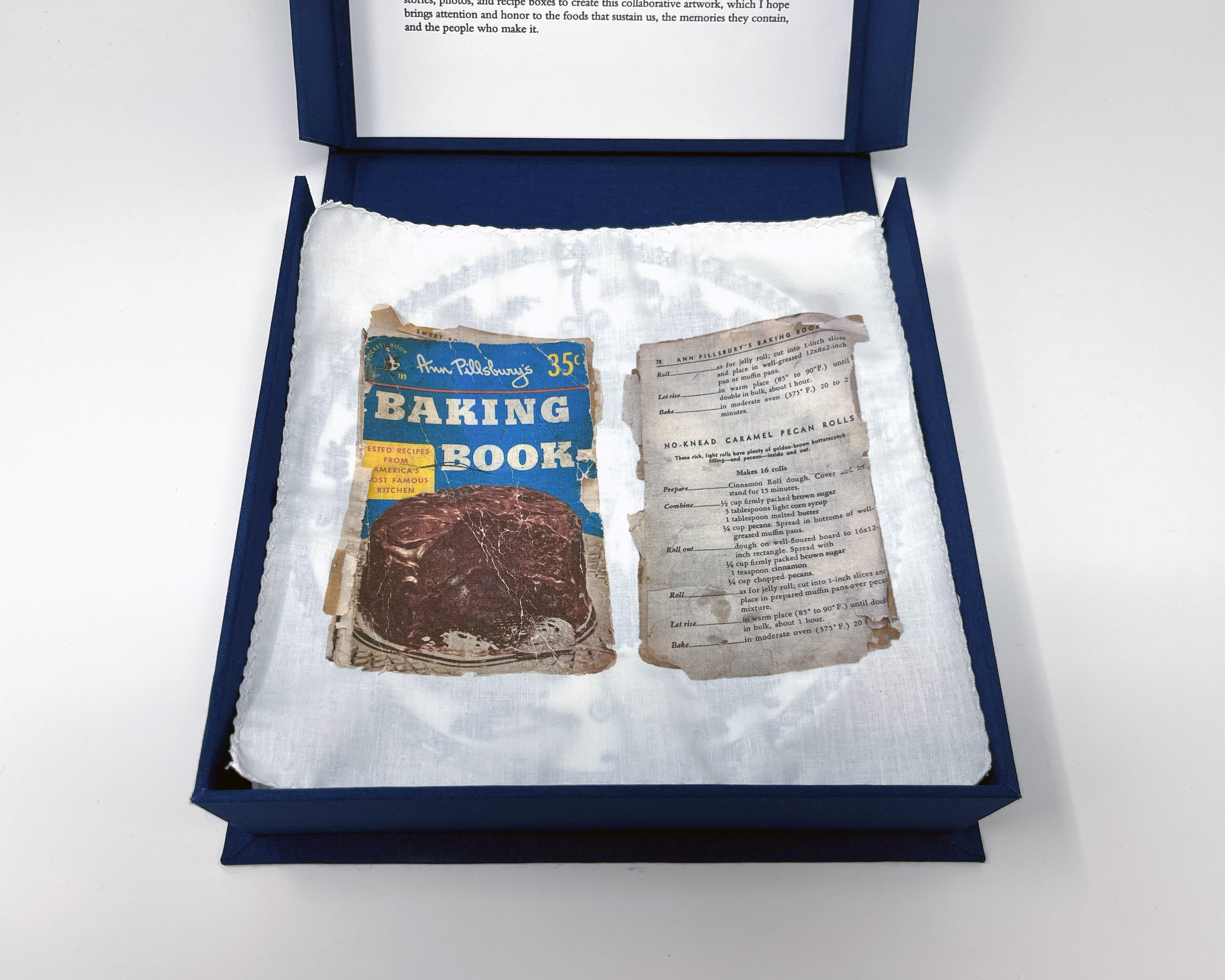 box open showing fabric liner underneath plate #3, printed with a photograph of the Ann Pillsbury's Baking Book and the page for "No-Knead Carmel Pecan Rolls" mentioned in Barbara's story previous.