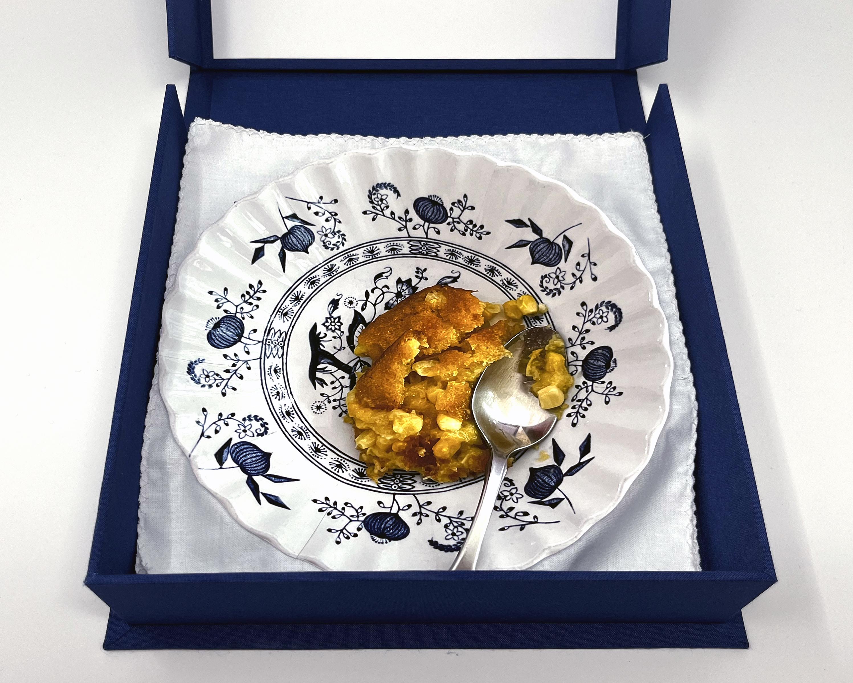 box open to show plate #6: a plate containing a photograph of corn casserole and a spoon on a blue patterned plate